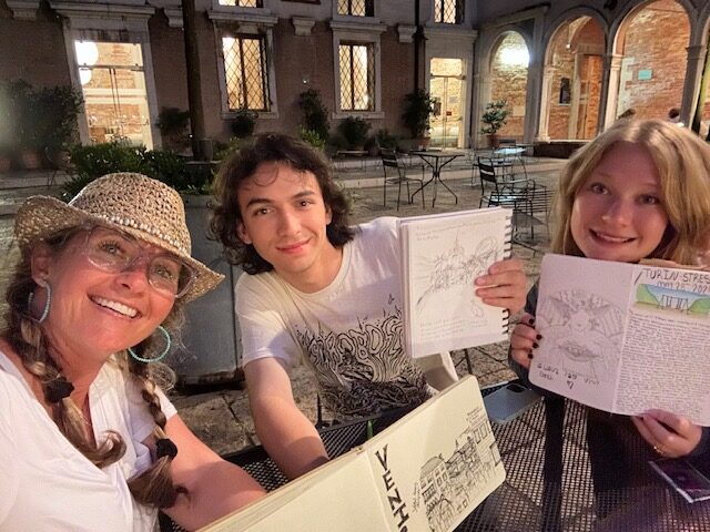 Three people sitting at an outdoor table, showing their drawings.