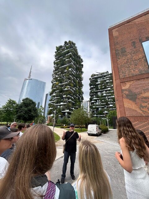 People gathered around a man speaking in front of two tall buildings covered in greenery, with a glass skyscraper and a brick building in the background.